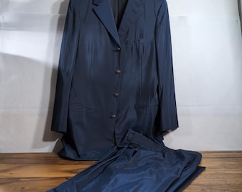 Può includere: Un completo blu scuro composto da una giacca monopetto a quattro bottoni e pantaloni abbinati. La giacca presenta revers classici, un taschino sul petto e due tasche con patta. Il tessuto presenta una sottile lucentezza uniforme.