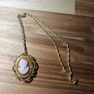May include: A gold-tone necklace featuring an oval pendant with a white profile cameo set against a terracotta-colored background. The pendant is framed by an ornate, textured gold-tone metal border and hangs from a twisted gold-tone chain.
