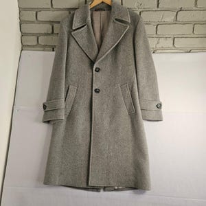 May include: A light gray wool coat with a classic design. The coat features a notched lapel, two front pockets, and button closures. The sleeves have buttoned cuffs. The coat is hanging on a wooden hanger against a white background.