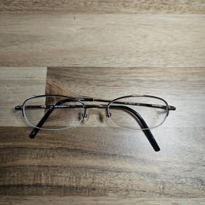 May include: Oval-shaped eyeglasses with a silver frame and black temples. The lenses are partially rimmed, and the glasses are resting on a wooden surface. The glasses have a classic, minimalist design.