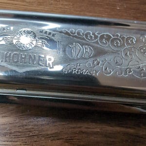 Könnte beinhalten: Eine silberfarbene Metall-Mundharmonika mit eingraviertem Hohner-Logo, dem Text GESETZL GESCH, einem Harfenemblem und dem Wort GERMANY. Die Oberfläche ist poliert und reflektierend und zeigt komplizierte Rankenmuster auf dem Metallgehäuse.