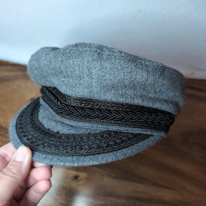 May include: A grey felted wool newsboy cap featuring a structured crown and a short, stiff brim. The cap is accented with a decorative black braided trim that wraps around the base of the crown and continues onto the edge of the brim.