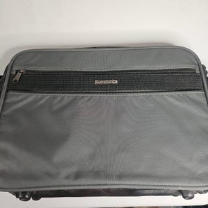 Puede incluir: Bolso para portátil Samsonite gris con cremallera negra y panel de malla negra. El logotipo de Samsonite se muestra en plata. El bolso tiene forma rectangular y parece estar hecho de un material duradero.