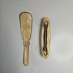 May include: Two antique vanity items. One is a long, paddle-shaped brush with a handle and a decorative leaf design. The other is a small, rectangular brush with a mirror. Both have a cream-colored, pearlescent finish with gold trim.