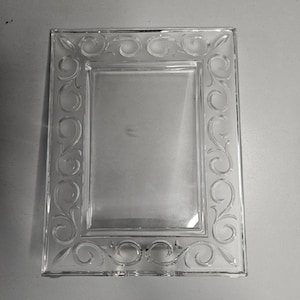 Puede incluir: Marco de fotos de vidrio transparente con diseño de volutas grabadas. El marco rectangular tiene un centro de vidrio transparente y detalles decorativos en los bordes. Diseñado para una fotografía o una obra de arte.