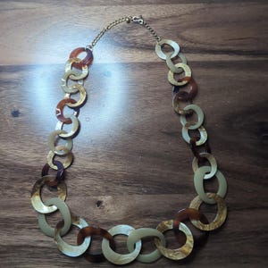 Può includere: Una collana caratterizzata da una serie di maglie circolari intrecciate in varie tonalità di crema, beige e marrone scuro. Le maglie hanno una finitura liscia e lucida e sono collegate nella parte superiore da una sottile catena in metallo dorato.