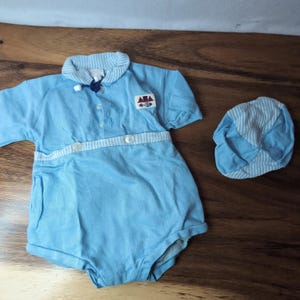 May include: A light blue long-sleeve romper with a white and blue striped collar and waist trim. The romper features a small patch with a red sailboat and anchor graphic on the chest, three white buttons, and a matching light blue and white striped cap.
