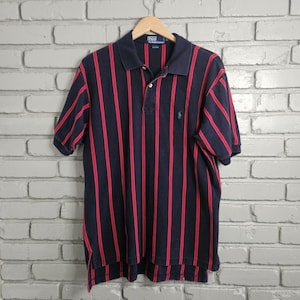 Könnte beinhalten: Marineblaues Poloshirt mit vertikalen roten Streifen. Das Shirt hat kurze Ärmel, einen Kragen und eine Knopfleiste mit drei Knöpfen. Ein kleines grünes Logo ist auf der linken Brust sichtbar. Das Shirt hängt an einem Holzbügel.