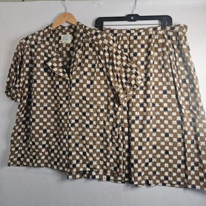 May include: A two-piece set consisting of a short-sleeved button-down shirt with a collar and a matching skirt. Both pieces feature a brown, white, and dark blue checkered pattern. The shirt has a visible label at the neckline and hangs on a hanger.