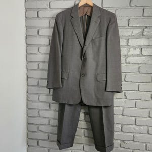 May include: A gray suit, consisting of a jacket and trousers, hangs against a white brick wall. The jacket has two buttons and a notched lapel. The trousers have a cuffed hem. The suit is made of a woven fabric.