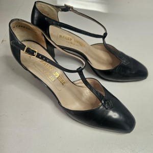 Può includere: Un paio di scarpe con tacco a T Bally Suisse in pelle nera. Le scarpe hanno la punta affusolata, un tacco piccolo e una chiusura con fibbia dorata. Il logo Bally Suisse è visibile sulla soletta.