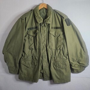 May include: An olive green field jacket featuring a fold-down collar, long sleeves, and four front pockets with buttoned flaps. The chest displays black embroidered text reading U.S. ARMY and MCCLELLAN, with a black patch on the left shoulder.