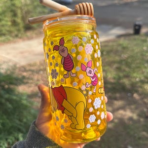 Puede incluir: Un tarro de miel de vidrio amarillo con un cucharón de miel de madera. El tarro está decorado con imágenes de dibujos animados de Winnie the Pooh y Piglet, junto con diseños florales blancos y rosas.