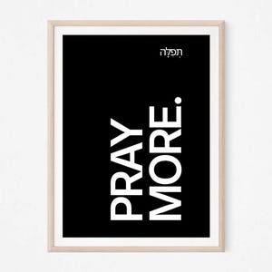 May include: A rectangular wall art print in a light wood frame featuring a solid black background with the white text PRAY MORE in a vertical orientation and the Hebrew word תפילה in white text positioned in the upper right corner of the print.