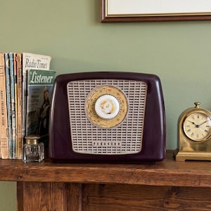 Op de afbeelding: Een vintage Ferguson buizenradio (c. 1953) met een donker bordeauxkleurige behuizing en een crèmekleurige wijzerplaat. De radio heeft een witte luidsprekergrille en staat op een houten plank met boeken en een kleine klok. De tekst "Radio Times" is zichtbaar.