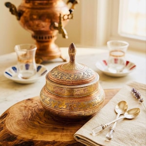 May include: An ornate brass container with a lid, featuring detailed etched patterns. The container sits on a wooden board, accompanied by tea glasses, saucers, and spoons, suggesting a tea ceremony. A copper samovar is visible in the background.