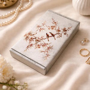 May include: A rectangular, silver-colored box with a detailed illustration of two birds perched on a branch with red and brown leaves. The box is set on a cream-colored fabric, with jewelry and flowers in the background.