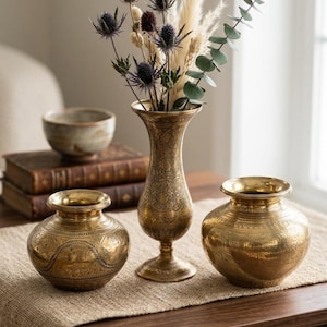 Può includere: Tre vasi antichi in ottone di varie dimensioni, con intricati disegni incisi, sono su un tavolo di legno. Il vaso più alto contiene un bouquet di fiori secchi e verde. I vasi sono esposti su un runner dai toni neutri.