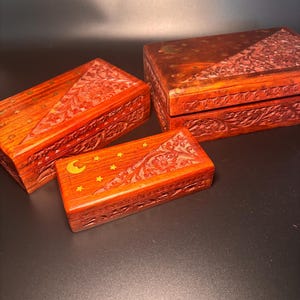 Könnte beinhalten: Drei rotbraune Holzkisten mit aufwendigen floralen Schnitzereien. Die kleinste Kiste zeigt ein Mond- und Stern-Design. Die rechteckigen Kisten sind unterschiedlich groß und eignen sich zur Aufbewahrung kleiner Gegenstände. Dekorativ.