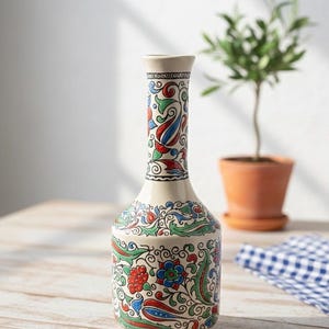 Puede incluir: Un jarrón de cerámica color crema con cuello delgado y base bulbosa. El jarrón está adornado con un vibrante patrón floral en rojo, azul y verde, sobre un fondo de líneas negras arremolinadas. Una pequeña etiqueta dice "Handmade in Greece - 1980s".