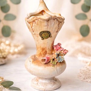 May include: Vintage Capodimonte vase. The handcrafted Italian porcelain vase has a bulbous base and a flared top. It features a cream and brown colour scheme with floral accents in pink, blue, and green. The vase has a decorative label.