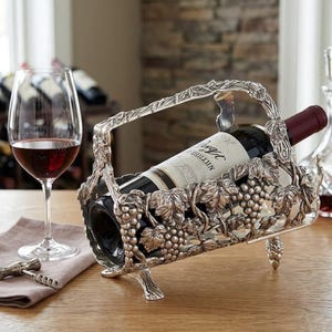 Puede incluir: Un soporte plateado para botellas de vino con asa, decorado con motivos de uvas y hojas. Una botella de vino está dentro del soporte. Una copa de vino tinto y un sacacorchos están sobre la mesa.