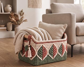 May include: A rectangular crochet storage basket featuring a geometric leaf pattern in sage green and terracotta yarn. The basket has a wooden handle and is filled with a beige knit throw blanket and a matching square throw pillow.