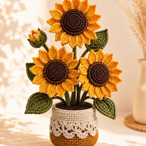 Könnte beinhalten: Ein gehäkeltes Sonnenblumenarrangement mit drei großen gelben Blüten mit dunkelbraunen Mitten, einer kleinen Knospe und grünen Blättern. Die Blumen stehen in einem zweifarbigen Topf mit einem cremefarbenen Oberteil und einer spitzenartigen Verzierung.
