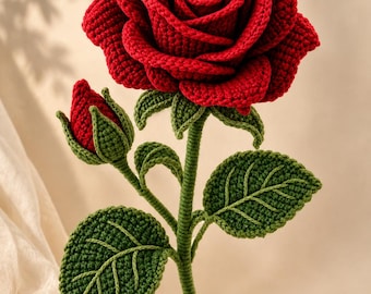 Puede incluir: Una rosa de ganchillo hecha a mano que presenta una gran flor de color rojo intenso con pétalos en capas, un capullo rojo cerrado más pequeño y hojas de color verde oscuro unidas a un tallo verde largo y delgado. La textura es visible.