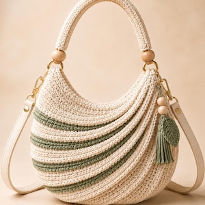 May include: A crescent-shaped crochet handbag in cream with sage green stripes. It features a rounded top handle with wooden beads, gold-tone metal hardware, a detachable shoulder strap, and a decorative sage green tassel with a leaf charm.