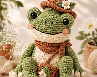 Puede incluir: Un peluche de rana tejido a ganchillo con cuerpo verde, vientre color crema, grandes ojos negros y mejillas rosadas. La rana lleva un sombrero marrón con una flor blanca, una bufanda marrón a juego y un pequeño bolso marrón con una flor.