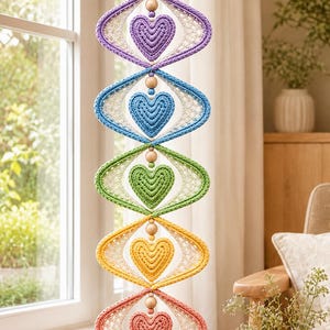 May include: A vertical macrame wall hanging featuring five stacked diamond-shaped frames in purple, blue, green, yellow, and coral. Each frame contains a matching colored heart-shaped macrame element, separated by small wooden beads.