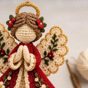 Pu&ograve; includere: Una statuetta di angelo all'uncinetto con capelli castani, un'aureola dorata e un vestito rosso. L'angelo ha ali color crema decorate con ricami di foglie verdi e bacche rosse. Il vestito presenta bordi color crema e piccoli fiori bianchi.