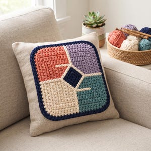 Op de afbeelding: Een vierkant sierkussen met een centraal gehaakt paneel met vier verschillende kleurblokken in koraal, lavendel, crème en groenblauw, gescheiden door crèmekleurige garenlijnen en omlijst door een donkerblauwe rand op een beige stoffen basis.