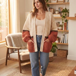 May include: An open-front crochet cardigan featuring a colour-blocked design with a cream upper section, a thin dark brown stripe, and a rust-coloured lower section. The garment includes long sleeves with ribbed cuffs, two front pockets, and a ribbed trim.