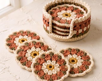 Puede incluir: Un juego de posavasos redondos de ganchillo con un patrón floral en tonos crema, terracota y verde oliva. Los posavasos están dispuestos en un soporte circular de ganchillo a juego con un borde festoneado, mostrando un tejido detallado.
