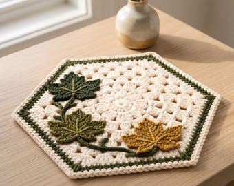 Puede incluir: Un tapete de ganchillo hexagonal con un patrón de granny square en color blanco roto y un borde verde oscuro. Tres hojas de arce texturizadas y tridimensionales en tonos verde oscuro, verde oliva y amarillo mostaza están fijadas.