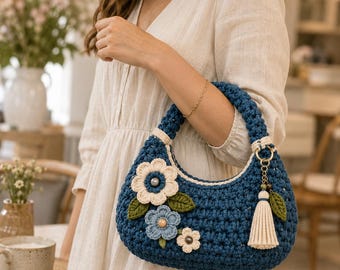 Puede incluir: Un bolso de ganchillo azul oscuro con forma de media luna y asa gruesa. El exterior está decorado con tres flores de ganchillo en color crema y azul claro, hojas verdes y un colgante de borla color crema unido a una anilla dorada.