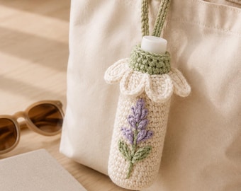 May include: A cream-colored crochet bottle cozy featuring a lavender sprig motif and a white daisy-shaped collar. The cozy has a sage green crochet handle and is attached to a plain canvas tote bag, showcasing intricate yarn work and textures.