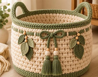 Puede incluir: Una cesta redonda de ganchillo color crema con un borde grueso verde salvia en la parte superior e inferior. La cesta está decorada con un lazo central de ganchillo verde salvia, dos borlas colgantes y dos motivos de hojas simétricos con cuentas de madera.