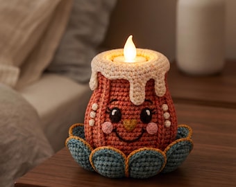 Puede incluir: Una funda de ganchillo para vela en color terracota con una cara sonriente, mejillas rosadas y un patrón de goteo color crema en la parte superior. La funda descansa sobre una base de ganchillo azul con forma de pétalos y contiene una vela LED.