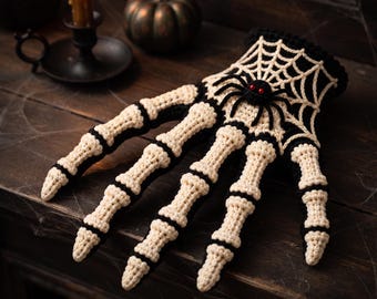 Puede incluir: Un guante de ganchillo diseñado para parecer una mano de esqueleto, con segmentos óseos de color blanco roto delineados en hilo negro. El dorso de la mano muestra un patrón de telaraña blanca detallada con una pequeña araña negra de ojos rojos.