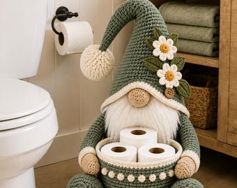 Puede incluir: Una figura de gnomo de ganchillo en color verde salvia con un sombrero alto y puntiagudo adornado con dos margaritas blancas y hojas verdes. El gnomo tiene una barba color crema, una nariz redonda y sostiene una cesta de ganchillo a juego con tres rollos de papel higiénico.