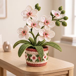 Könnte beinhalten: Eine gehäkelte Orchidee mit weißen Blütenblättern, rosa Zentren und grünen Knospen, in einem runden Topf mit einem dekorativen Band aus kleinen weißen Blüten und grünen Blättern, abgerundet durch eine strukturierte rosa-cremefarbene Basis.
