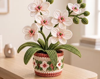 Puede incluir: Una orquídea de ganchillo con pétalos blancos, centros rosados y capullos verdes, plantada en un recipiente redondo con una banda decorativa de pequeñas flores blancas y hojas verdes, con una base de ganchillo en tonos rosa y crema.
