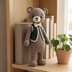May include: A brown crocheted teddy bear toy featuring a dark green vest with cream-colored trim and a matching cream-colored bow tie. The bear has dark eyes, a stitched nose, and rounded ears with cream-colored centers, standing upright.