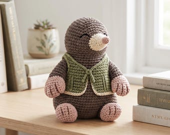 Puede incluir: Un peluche de topo tejido a ganchillo en color marrón que lleva un chaleco verde con ribete color crema. El topo tiene un hocico color crema, patas y pies de color rosa tostado y ojos cerrados bordados. La figura tiene puntos visibles.