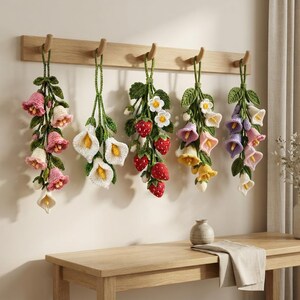 May include: Five hanging crochet wall decorations featuring botanical themes. The arrangements include pink and cream bell-shaped flowers, white calla lilies, red strawberries with white blossoms, yellow and purple bell flowers, and green leaves.