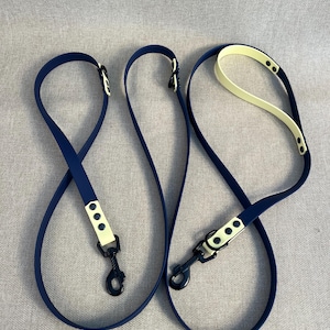 Könnte beinhalten: Zwei marineblaue Hundeleinen mit cremefarbenen Akzenten und schwarzen Metallverschlüssen. Die Leinen bestehen aus einem flexiblen Material und sind für Tierhalter konzipiert. Der Hintergrund ist ein neutraler grauer Stoff.