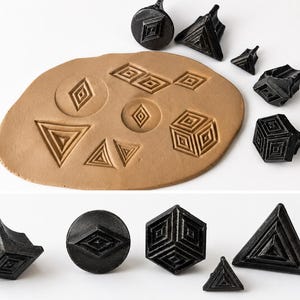 May include: A collection of black geometric stamps with various shapes, including diamonds, triangles, and cubes. Some stamps are pressed into a tan clay surface, showcasing the imprinted designs. The stamps are arranged on a white background.